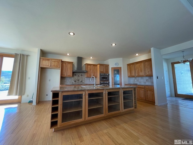 kitchen featuring wall chimney range hood, backsplash, a kitchen island with sink, light hardwood / wood-style floors, and sink