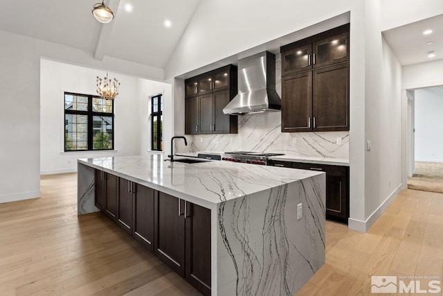 kitchen featuring light stone counters, an island with sink, wall chimney range hood, light wood-type flooring, and vaulted ceiling