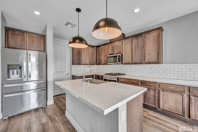 kitchen with sink, appliances with stainless steel finishes, an island with sink, light hardwood / wood-style flooring, and pendant lighting