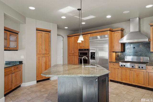 kitchen with appliances with stainless steel finishes, decorative light fixtures, wall chimney exhaust hood, and a kitchen island with sink