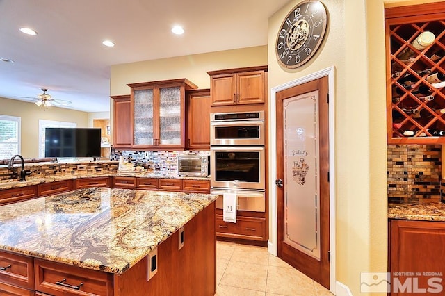 kitchen featuring light tile patterned flooring, backsplash, light stone countertops, double oven, and ceiling fan