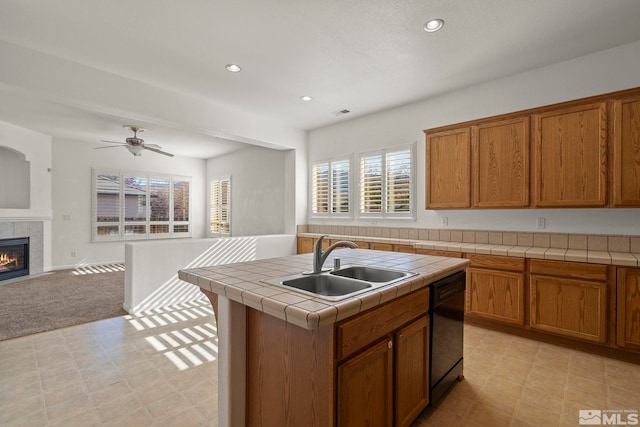 kitchen featuring a center island with sink, sink, tile counters, a tile fireplace, and black dishwasher