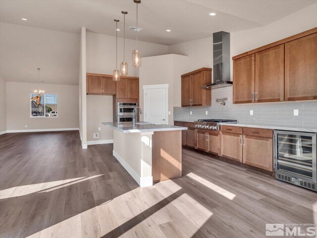 kitchen featuring beverage cooler, hanging light fixtures, a kitchen island with sink, wall chimney range hood, and wood-type flooring