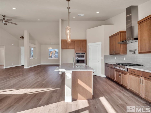 kitchen with stainless steel appliances, wall chimney range hood, an island with sink, decorative light fixtures, and light wood-type flooring