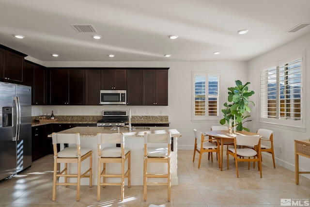 kitchen featuring a kitchen bar, an island with sink, dark brown cabinets, light stone countertops, and stainless steel appliances