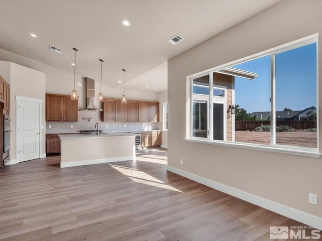 kitchen featuring a kitchen island with sink, wall chimney range hood, light hardwood / wood-style flooring, pendant lighting, and decorative backsplash