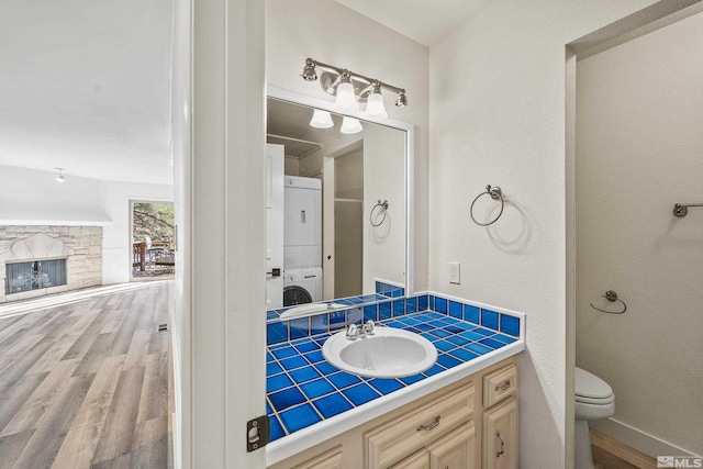 bathroom featuring vanity, wood-type flooring, stacked washer and clothes dryer, toilet, and a stone fireplace