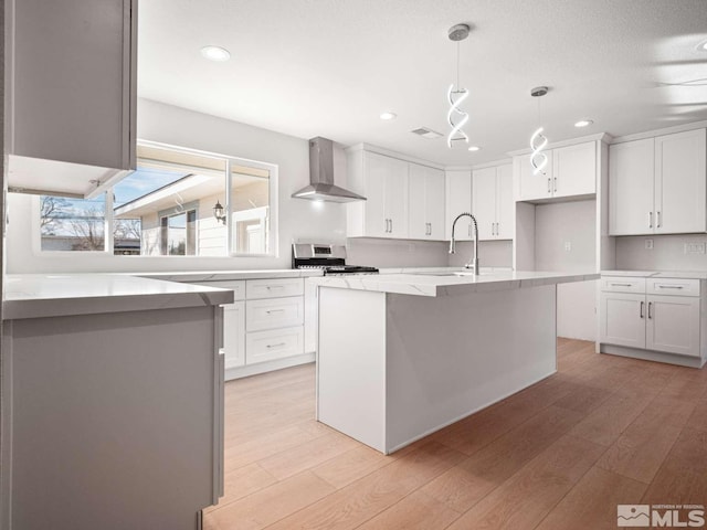 kitchen with light wood-type flooring, stainless steel gas range oven, wall chimney exhaust hood, white cabinets, and hanging light fixtures