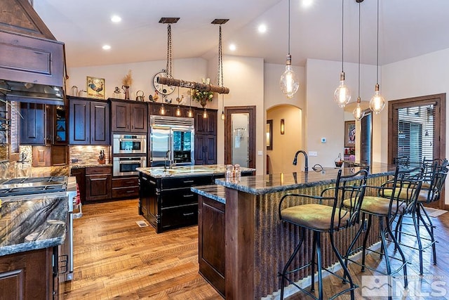 kitchen with high quality appliances, backsplash, a center island with sink, light wood-type flooring, and dark brown cabinetry