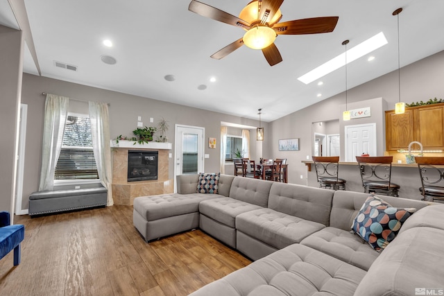 living room with a tiled fireplace, ceiling fan, vaulted ceiling with skylight, and light hardwood / wood-style floors