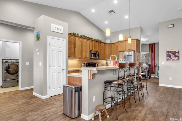 kitchen featuring hanging light fixtures, an island with sink, washer / dryer, wood-type flooring, and stainless steel appliances