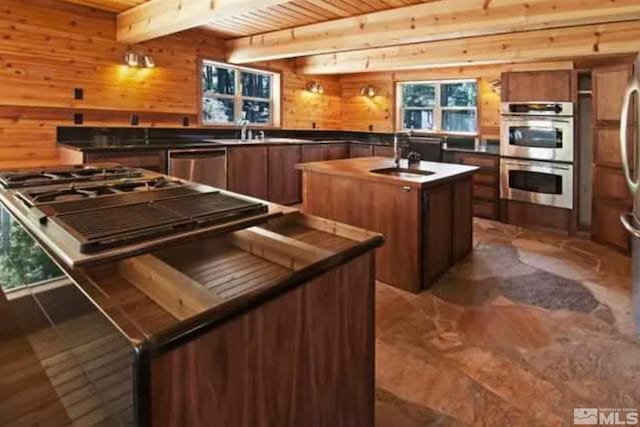 kitchen with sink, beamed ceiling, wood walls, a kitchen island with sink, and appliances with stainless steel finishes