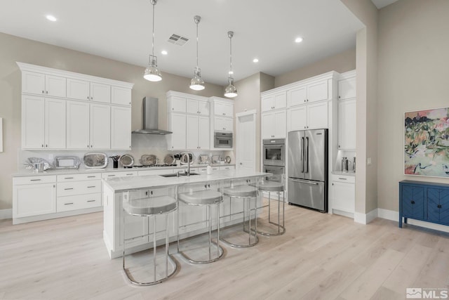 kitchen with wall chimney exhaust hood, hanging light fixtures, stainless steel appliances, a center island with sink, and white cabinets