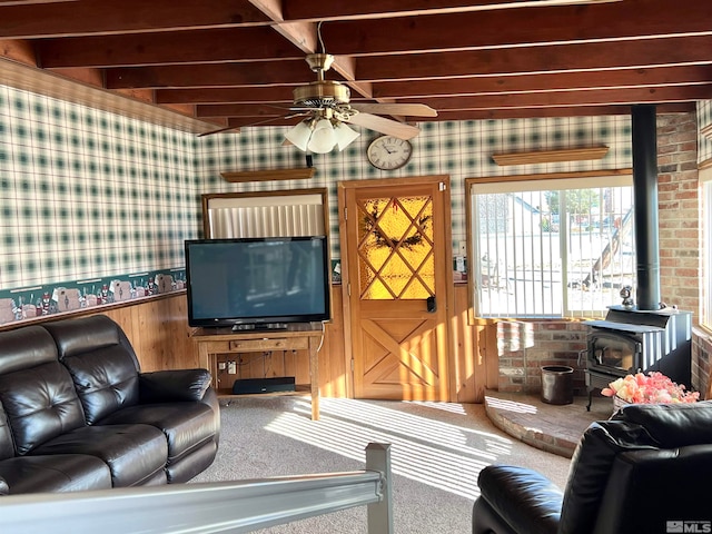 carpeted living room with a wood stove, ceiling fan, beamed ceiling, brick wall, and wooden walls