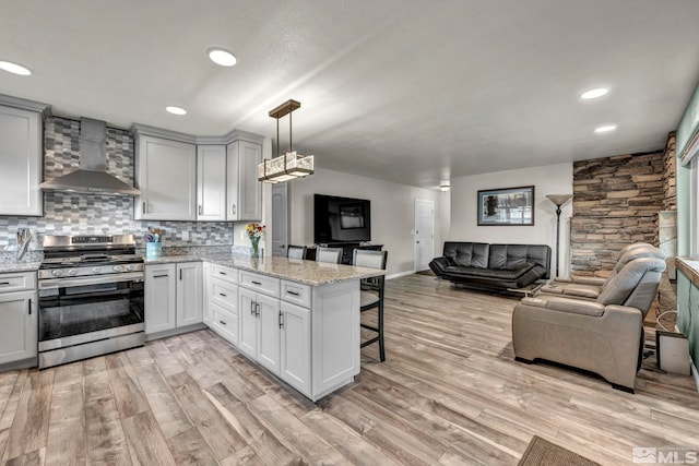 kitchen featuring wall chimney range hood, stainless steel stove, light wood-type flooring, decorative light fixtures, and kitchen peninsula