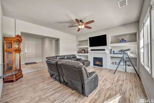 living room featuring ceiling fan, light wood-type flooring, built in shelves, and a tiled fireplace