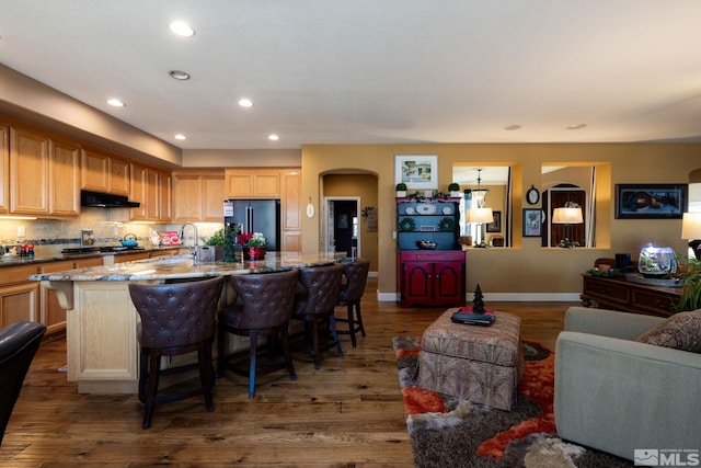 kitchen with stone counters, dark hardwood / wood-style flooring, light brown cabinetry, a kitchen island with sink, and black refrigerator