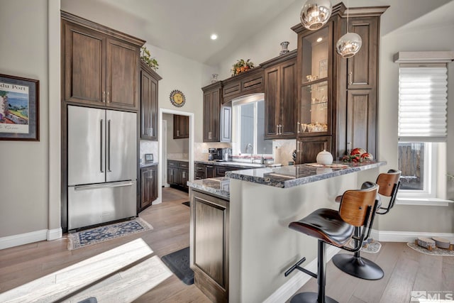 kitchen with light wood-type flooring, stainless steel built in refrigerator, dark brown cabinetry, dark stone countertops, and lofted ceiling