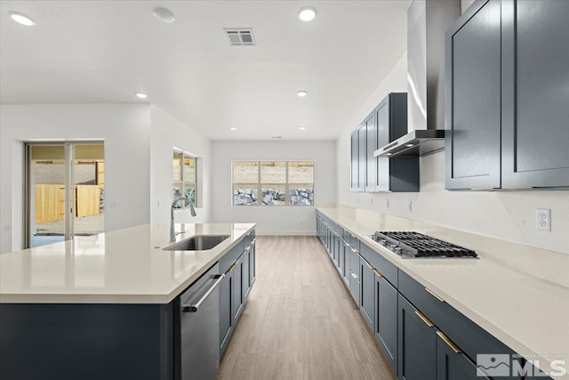 kitchen featuring light wood-type flooring, stainless steel appliances, a kitchen island with sink, wall chimney range hood, and sink