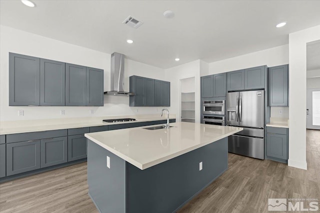 kitchen with wall chimney exhaust hood, a kitchen island with sink, stainless steel fridge with ice dispenser, sink, and hardwood / wood-style floors