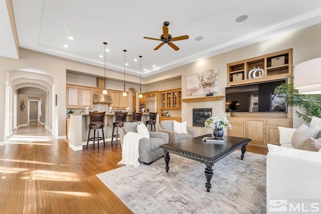 living room with a tile fireplace, ceiling fan, built in shelves, light wood-type flooring, and a tray ceiling