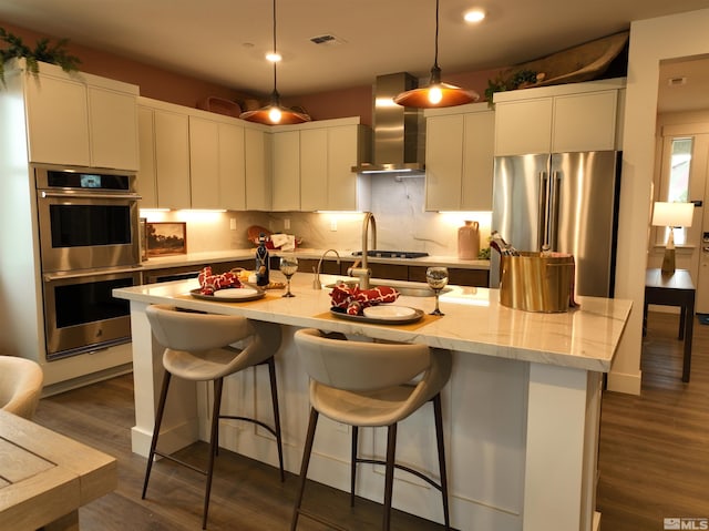 kitchen with an island with sink, appliances with stainless steel finishes, hanging light fixtures, wall chimney range hood, and white cabinetry