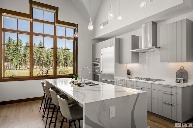 kitchen with appliances with stainless steel finishes, wall chimney exhaust hood, a kitchen island with sink, sink, and decorative light fixtures