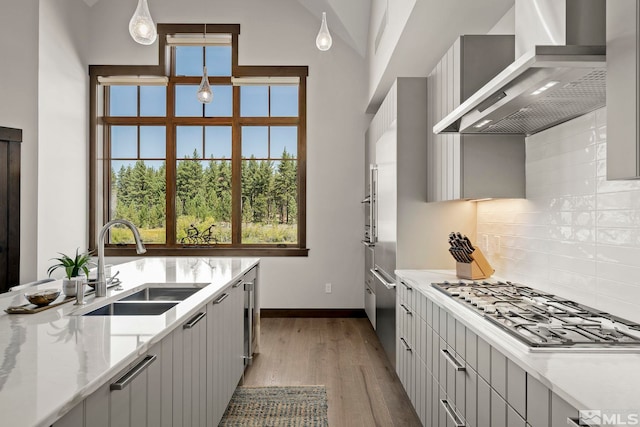 kitchen with sink, wall chimney exhaust hood, hanging light fixtures, light stone counters, and light hardwood / wood-style flooring