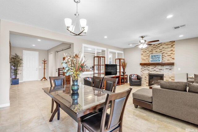 tiled dining space with ceiling fan with notable chandelier and a stone fireplace