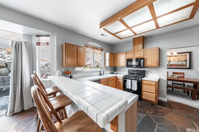 kitchen with tile counters, stone tile flooring, a sink, a peninsula, and black appliances