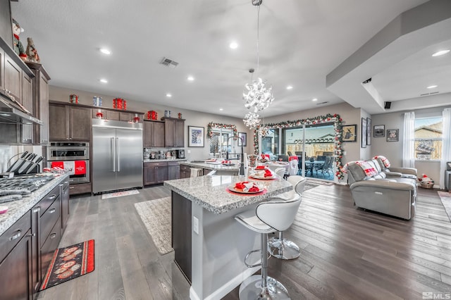 kitchen featuring dark brown cabinetry, light stone countertops, an island with sink, decorative light fixtures, and appliances with stainless steel finishes