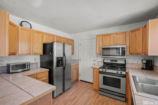 kitchen with sink, tile counters, stainless steel appliances, light hardwood / wood-style flooring, and light brown cabinetry