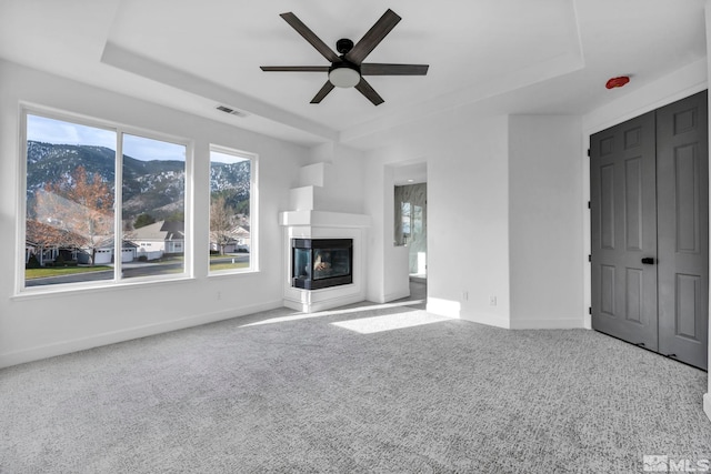 unfurnished living room with carpet flooring, ceiling fan, a multi sided fireplace, a raised ceiling, and a mountain view