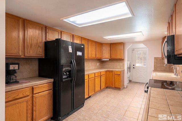 kitchen featuring washer and clothes dryer, tile countertops, black refrigerator with ice dispenser, and tasteful backsplash