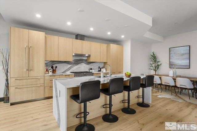 kitchen with sink, light wood-type flooring, an island with sink, light brown cabinetry, and tasteful backsplash