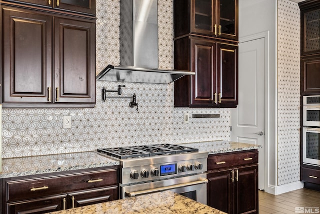kitchen featuring hardwood / wood-style flooring, wall chimney exhaust hood, appliances with stainless steel finishes, dark brown cabinets, and light stone counters