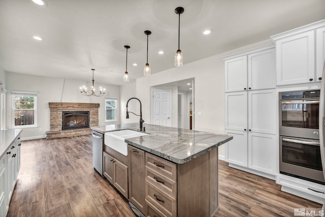 kitchen with light stone countertops, appliances with stainless steel finishes, white cabinets, hanging light fixtures, and an island with sink