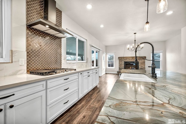 kitchen with sink, wall chimney exhaust hood, hanging light fixtures, a stone fireplace, and white cabinets