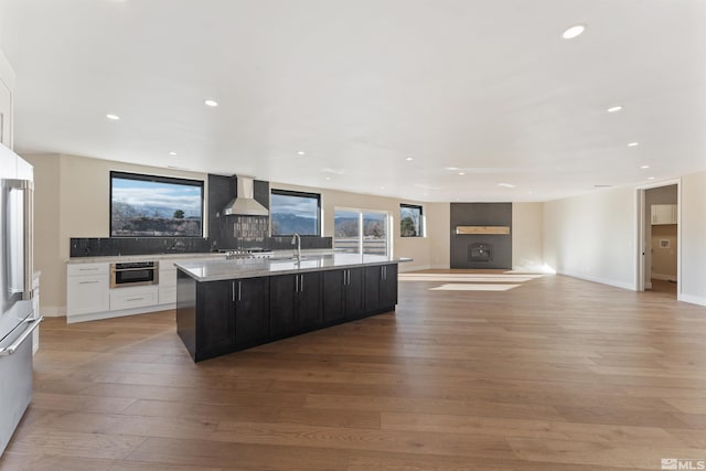 kitchen with a center island with sink, light wood-type flooring, wall chimney range hood, and tasteful backsplash