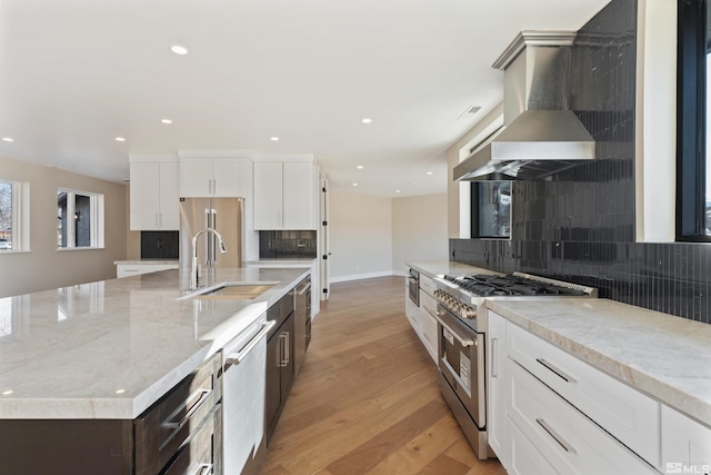 kitchen featuring white cabinetry, light stone countertops, wall chimney range hood, a large island with sink, and high end appliances