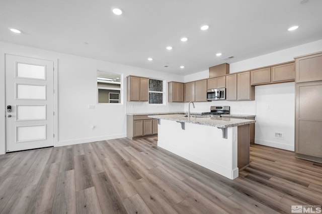 kitchen with appliances with stainless steel finishes, a kitchen island with sink, light stone counters, and light hardwood / wood-style floors