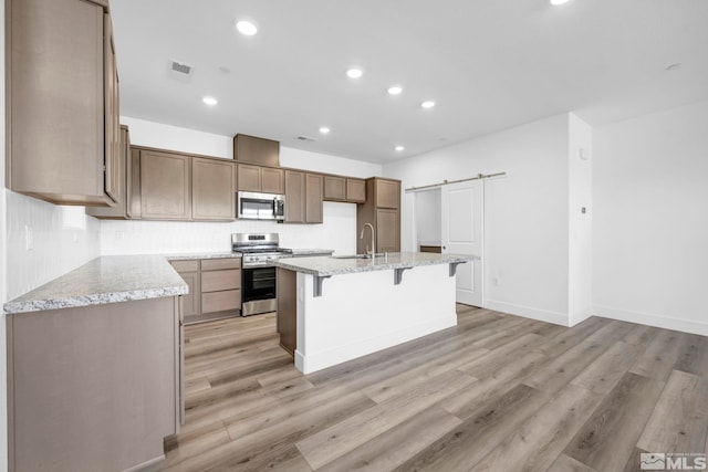 kitchen with sink, light stone counters, appliances with stainless steel finishes, an island with sink, and a barn door