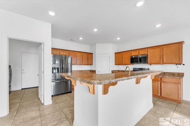 kitchen with light tile patterned floors, an island with sink, appliances with stainless steel finishes, a kitchen bar, and washer / clothes dryer