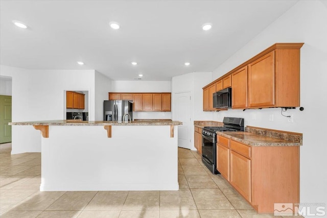 kitchen featuring a breakfast bar, black appliances, light stone countertops, light tile patterned floors, and an island with sink