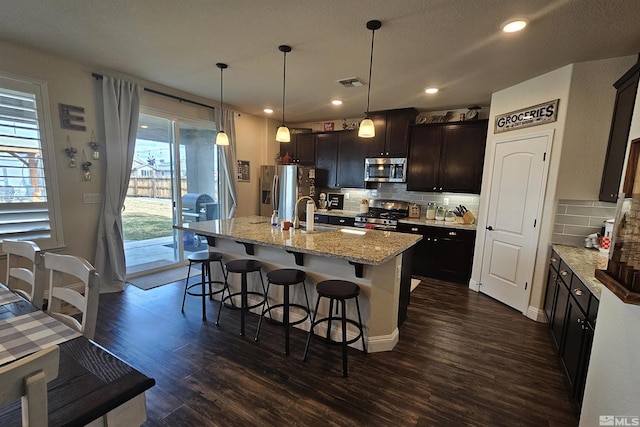 kitchen with pendant lighting, backsplash, dark brown cabinetry, a kitchen island with sink, and appliances with stainless steel finishes