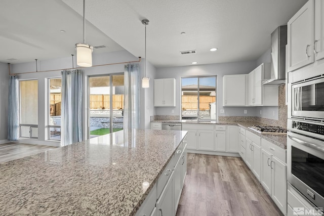 kitchen featuring appliances with stainless steel finishes, light wood-type flooring, light stone counters, wall chimney exhaust hood, and white cabinetry