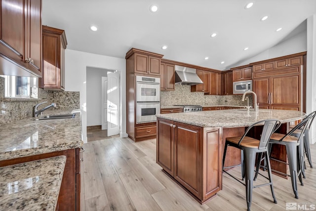 kitchen featuring light stone countertops, wall chimney range hood, an island with sink, vaulted ceiling, and appliances with stainless steel finishes