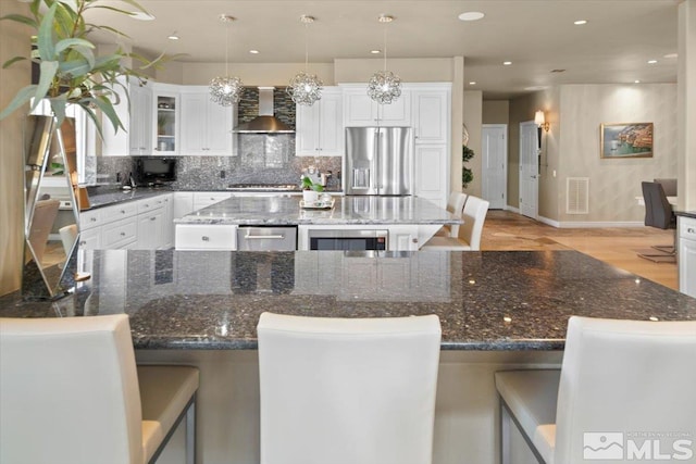 kitchen featuring white cabinets, wall chimney range hood, appliances with stainless steel finishes, decorative light fixtures, and a kitchen island