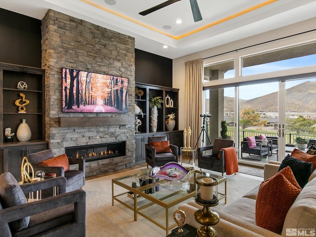 living room with a tray ceiling, a stone fireplace, and ceiling fan