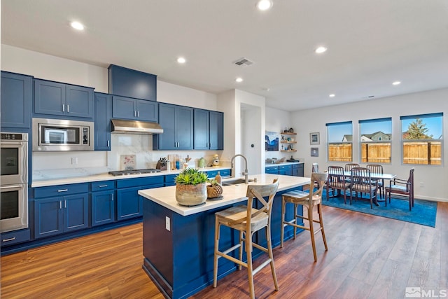 kitchen with appliances with stainless steel finishes, a breakfast bar, blue cabinetry, a center island with sink, and dark hardwood / wood-style floors